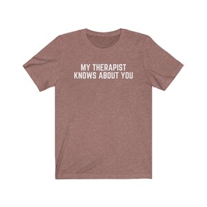 May include: A heather brown t-shirt with the text "MY THERAPIST KNOWS ABOUT YOU" printed in white.