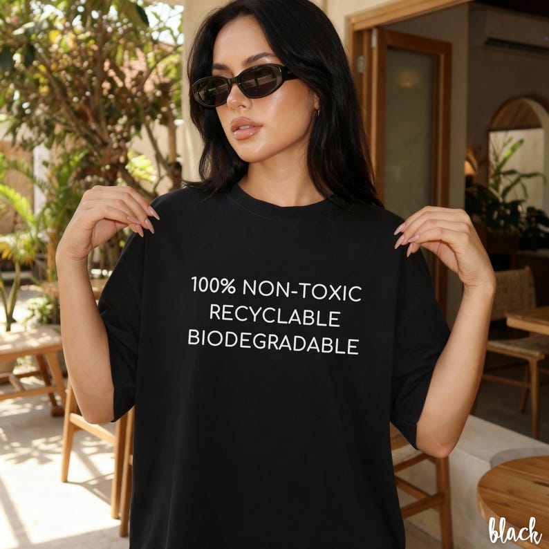 May include: Black t-shirt with white text: "100% NON-TOXIC, RECYCLABLE, BIODEGRADABLE." The shirt is a solid black color. The model is wearing sunglasses. The shirt is a casual style.