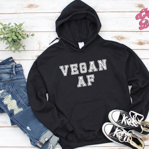 Op de afbeelding: Zwarte hoodie met de tekst "VEGAN AF" in witte blokletters. De hoodie heeft een voorzak en een capuchon met trekkoord. Een paar zwart-witte sneakers en een blauwe jeans liggen naast de hoodie.