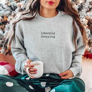 May include: A heather gray sweatshirt with the text "literally freezing" in black. The person is holding a white mug. The person is wearing green and white polka dot pajama pants. The background includes a Christmas tree.