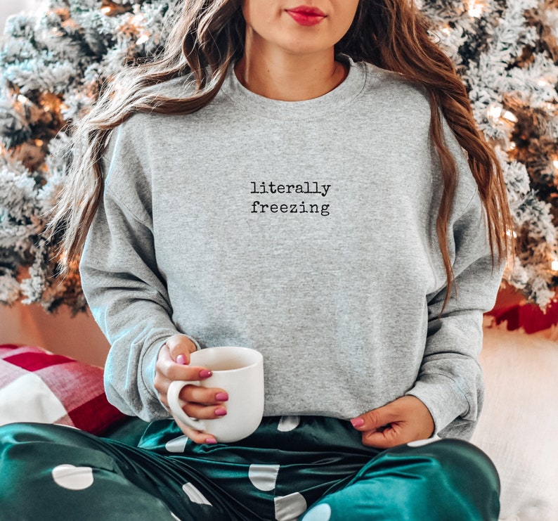 May include: A heather gray sweatshirt with the text "literally freezing" in black. The person is holding a white mug. The person is wearing green and white polka dot pajama pants. The background includes a Christmas tree.