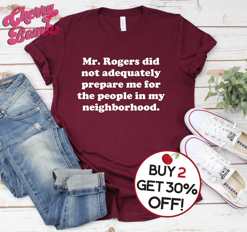 Puede incluir: Una camiseta color burdeos con texto blanco que dice "Mr. Rogers no me prepar&oacute; adecuadamente para la gente de mi vecindario." Un c&iacute;rculo rojo con una cereza blanca y el texto "COMPRA 2, LLEVA 30% DE DESCUENTO!" se encuentra en la esquina inferior derecha de la imagen.