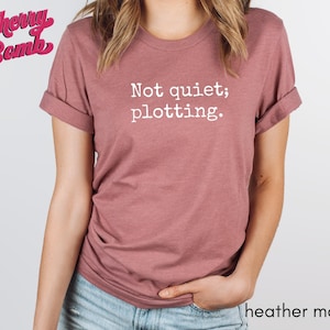 May include: Heather mauve t-shirt with rolled sleeves. The shirt features the text "Not quiet; plotting." in white. The Cherry Bomb logo is in the upper left corner. The words "heather mauve" are at the bottom right.