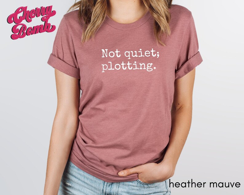 May include: Heather mauve t-shirt with rolled sleeves. The shirt features the text "Not quiet; plotting." in white. The Cherry Bomb logo is in the upper left corner. The words "heather mauve" are at the bottom right.