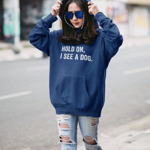 Pode incluir: Uma mulher vestindo um moletom azul marinho com o texto "HOLD ON, I SEE A DOG." em letras brancas. Ela est&aacute; vestindo jeans azul com rasgos e buracos.