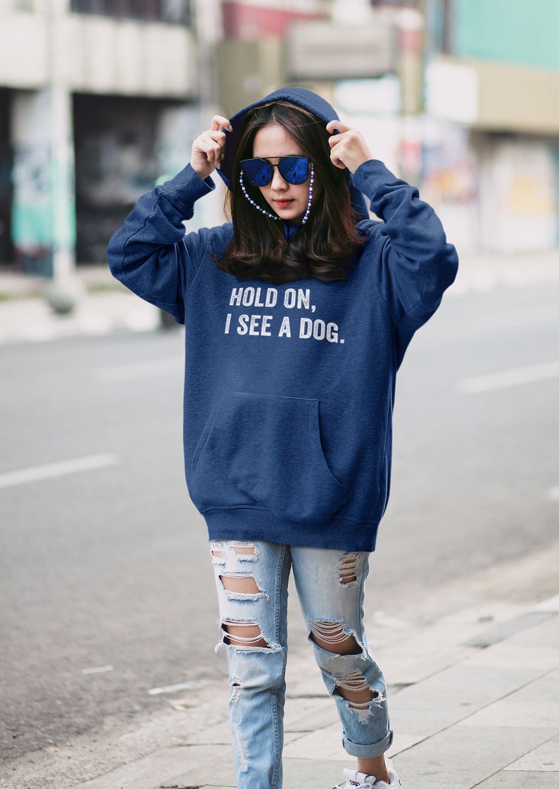 Pode incluir: Uma mulher vestindo um moletom azul marinho com o texto "HOLD ON, I SEE A DOG." em letras brancas. Ela est&aacute; vestindo jeans azul com rasgos e buracos.
