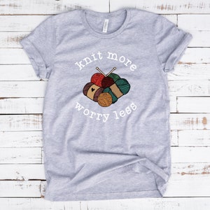 May include: A light gray t-shirt with the words "knit more worry less" in white script. The shirt features a graphic of yarn balls in various colors, including red, brown, and teal, with knitting needles.