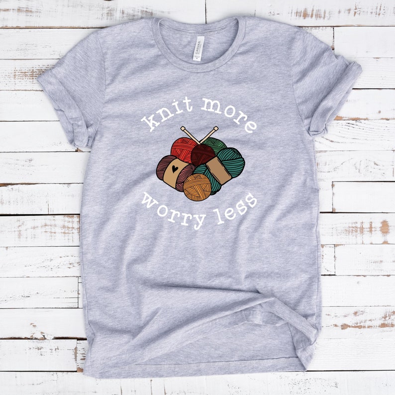 May include: A light gray t-shirt with the words "knit more worry less" in white script. The shirt features a graphic of yarn balls in various colors, including red, brown, and teal, with knitting needles.