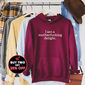 May include: Burgundy hoodie with a white print that says "I am a motherfucking delight."  The hoodie is hanging on a rack with other clothing. There is a red and white sticker that says "Buy Two Get 25% Off" in the lower left corner of the image.