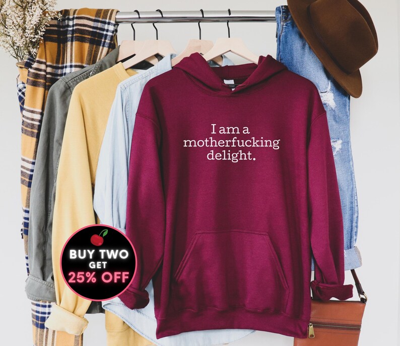 May include: Burgundy hoodie with a white print that says "I am a motherfucking delight."  The hoodie is hanging on a rack with other clothing. There is a red and white sticker that says "Buy Two Get 25% Off" in the lower left corner of the image.