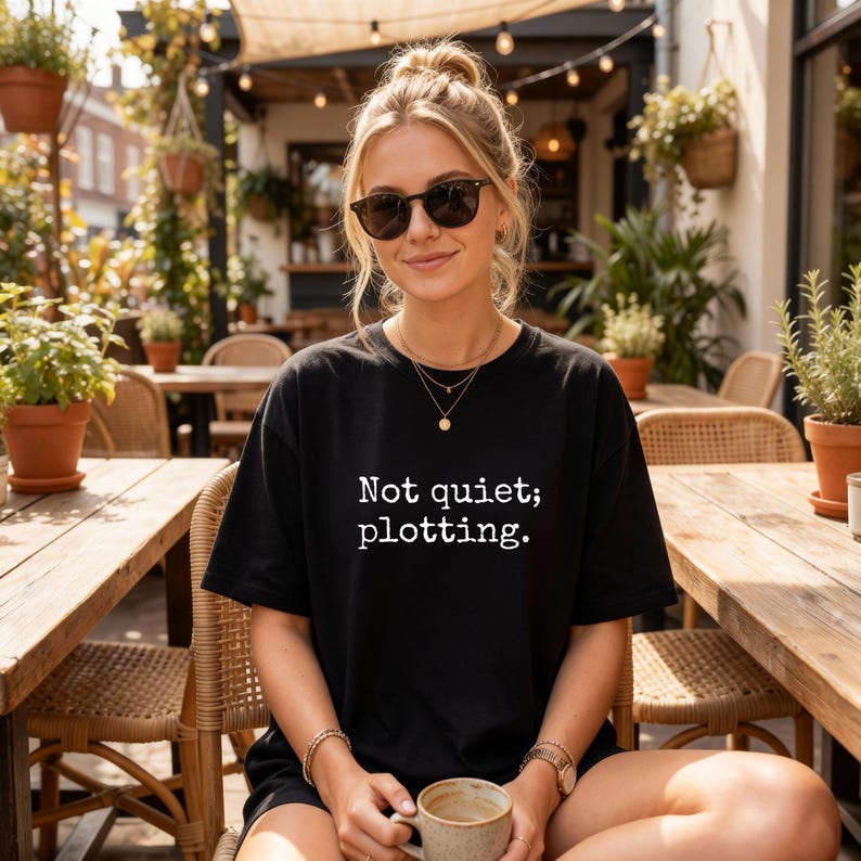 May include: A black t-shirt with the text "Not quiet; plotting." in white. The person is wearing sunglasses and a gold necklace, sitting at a wooden table outdoors. The person is holding a mug.