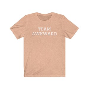 May include: A light peach colored t-shirt with the text "TEAM AWKWARD" printed in white on the front.