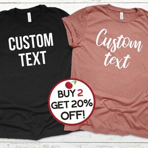 May include: Two t-shirts, one black and one dusty rose, each with the words "CUSTOM TEXT" printed in white. A circular graphic with a cherry and the text "BUY 2 GET 20% OFF!" is in the foreground.