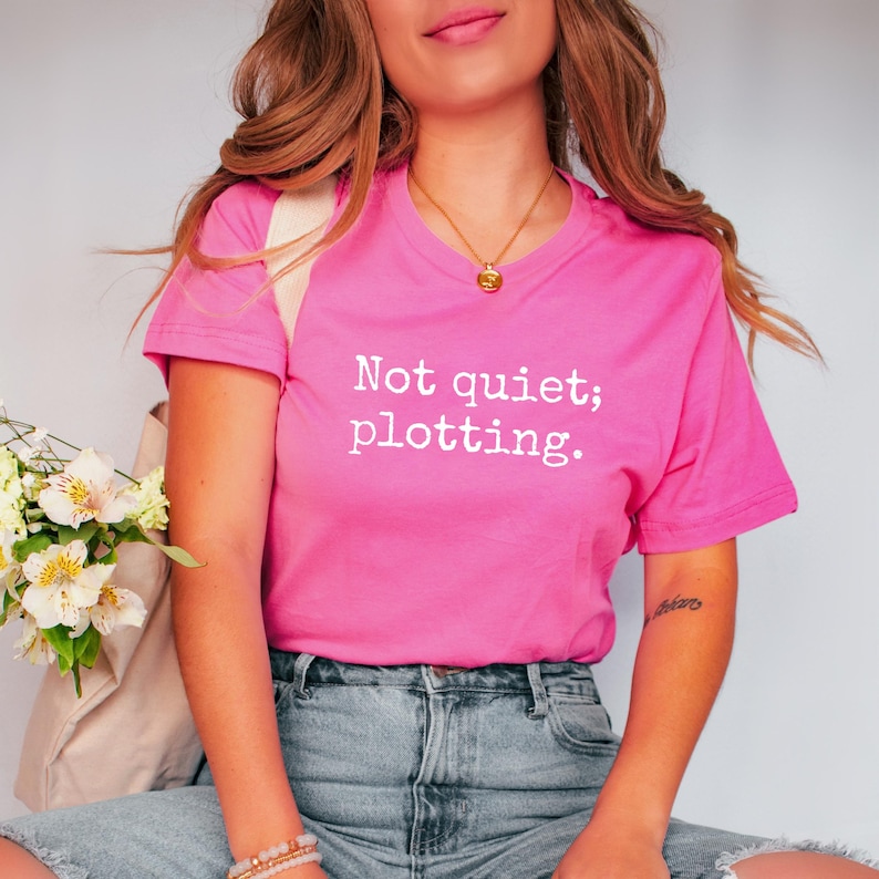 May include: A bright pink t-shirt with the text "Not quiet; plotting." in white. The shirt is a crew neck style and is paired with blue denim jeans. The overall aesthetic is casual and comfortable.