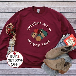 May include: A maroon sweatshirt with the text "crochet more worry less" and a graphic of yarn balls and a crochet hook. The text "BUY 2 GET 30% OFF!" is on a red circle sticker.
