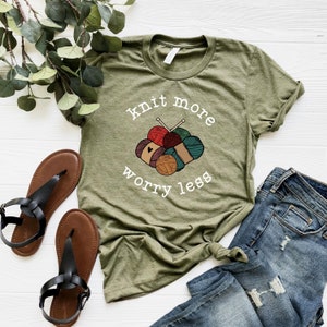 May include: Olive green t-shirt with the text "knit more worry less" above a graphic of yarn balls and knitting needles. The shirt is paired with blue jeans and black sandals.
