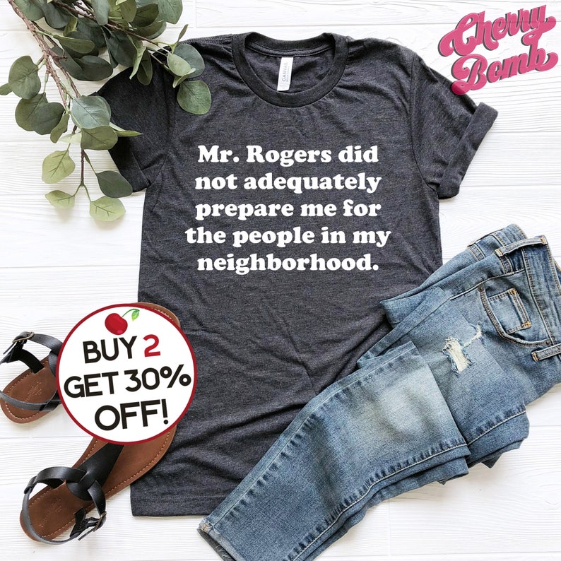 Puede incluir: Una camiseta gris oscuro con texto blanco que dice "Mr. Rogers did not adequately prepare me for the people in my neighborhood." La camiseta est&aacute; doblada y se muestra sobre una superficie blanca con un par de jeans azules y un gr&aacute;fico de cereza roja con el texto "BUY 2 GET 30% OFF!"