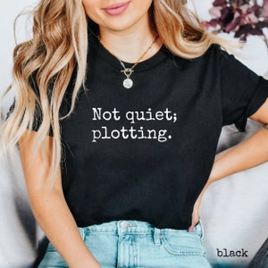 May include: Black crew neck t-shirt with the white text "Not quiet; plotting." printed on the front. The shirt is a solid black color and is made of a soft material. The word "black" is printed at the bottom right.
