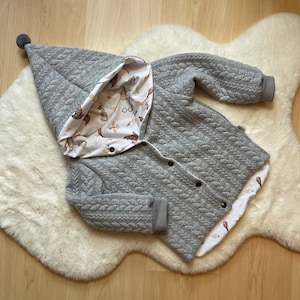 Könnte beinhalten: Eine graue Kapuzenjacke mit Steppmuster und einem weißen Futter mit einem verspielten Druck von Sternen, Brillen und anderen magischen Objekten. Die Jacke hat einen Knopfverschluss und einen Bommel an der Kapuze.