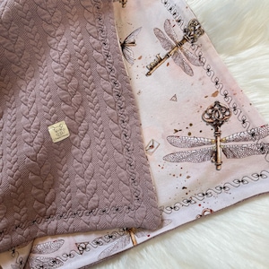 Könnte beinhalten: Eine mauvefarbene gesteppte Decke mit einem dekorativen Rand. Die Decke ist so gefaltet, dass ein rosa-weißes Stoff mit einem Muster aus Libellenflügeln und Schlüsseln sichtbar wird.