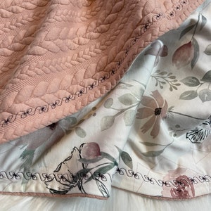 Könnte beinhalten: Eine rosafarbene gesteppte Decke mit einem Blumenmuster auf der Rückseite. Die Decke hat einen dekorativen Stich an den Rändern.