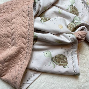 Könnte beinhalten: Eine rosa-weiße Babydecke mit gesteppter Textur und einem Muster aus grünen und braunen Schildkröten auf weißem Hintergrund. Die Decke hat einen dekorativen Rand.