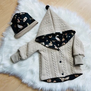 Könnte beinhalten: Eine beige Kapuzenjacke mit einem Zopfmuster und einem schwarzen und weißen Eulen-Print-Futter. Die Jacke hat einen schwarzen Bommel an der Kapuze und einen Knopfverschluss. Eine passende schwarze und weiße Eulen-Print-Mütze ist im Lieferumfang enthalten.