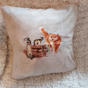 Könnte beinhalten: Beiges Zierkissen mit einer Aquarellillustration einer orangefarbenen Katze, einer grauen Ratte, einem Bücherstapel und einer Laterne. Die Katze steht, der Schwanz ist erhoben. Die Ratte befindet sich auf den Büchern.
