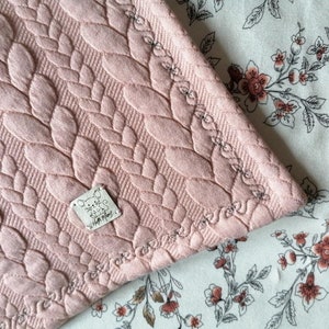 Könnte beinhalten: Eine hellrosa gesteppte Decke mit einem Blättermuster. Die Decke hat einen dekorativen, genähten Rand und ein kleines Etikett mit Text und einer Cartoon-Maus. Die Decke bedeckt teilweise einen Stoff mit floralem Muster.