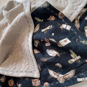 Könnte beinhalten: Eine beige gesteppte Decke mit einem schwarzen und weißen Eulen-Druck. Die Decke hat einen dekorativen Stich an den Rändern. Der Eulen-Druck zeigt Eulen, Schriftrollen und andere magische Gegenstände.