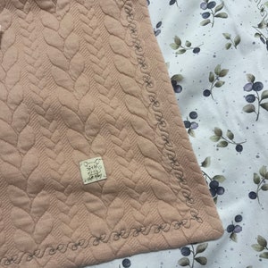Könnte beinhalten: Eine hellrosa gesteppte Babydecke mit einem dekorativen, gesteppten Rand. Die Decke hat ein kleines weißes Etikett mit dem Text "Little Mummy" darauf.