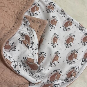 Könnte beinhalten: Eine rosafarbene gesteppte Babydecke mit einem weißen Baumwollfutter, das ein sich wiederholendes Muster aus schlafenden braunen Füchsen in einem Kreis aus Blättern zeigt. Die Decke hat einen dekorativen Rand aus schwarzen Nähten.