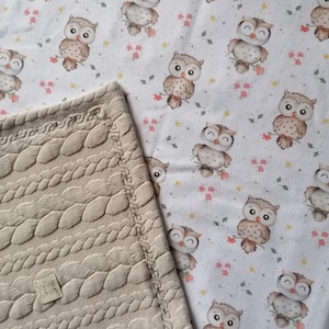Könnte beinhalten: Eine beige gestrickte Babydecke mit einem Zopfmuster. Die Decke ist in der Hälfte gefaltet und liegt auf einem weißen Stoff mit einem sich wiederholenden Muster aus braunen Eulen mit rosa Blumen.