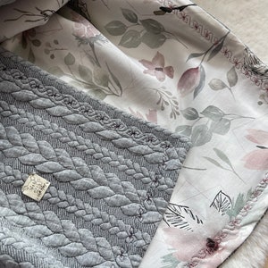 Könnte beinhalten: Eine graue gesteppte Decke mit einem floralen Muster auf der Rückseite. Die Decke hat eine gewebte Textur und ein zartes rosafarbenes Blumenmuster. Die Decke ist in der Hälfte gefaltet und die Blumen Seite ist sichtbar.