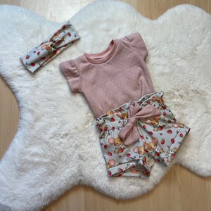 Könnte beinhalten: Ein rosa-weißes Baby-Outfit mit einem kurzärmeligen Oberteil und einer Shorts. Die Shorts haben einen Mäuse- und Erdbeer-Druck. Das Oberteil hat ein Punkte-Muster. Das Outfit liegt auf einem weißen Kunstfell-Teppich.