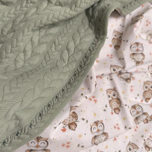 Könnte beinhalten: Eine salbeigrüne Steppdecke mit Blättermuster ist über einem weißen Stoff drapiert, der mit Cartoon-Eulen und kleinen Blumen bedruckt ist. Der Rand der Decke ist mit einer dekorativen Naht versehen.