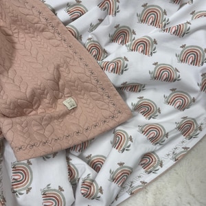 Könnte beinhalten: Eine rosafarbene gesteppte Babydecke mit einem weißen Baumwollstoff, der ein sich wiederholendes Muster aus mehrfarbigen Regenbögen mit Schmetterlingen zeigt. Die Decke ist in der Mitte gefaltet und zeigt beide Seiten.