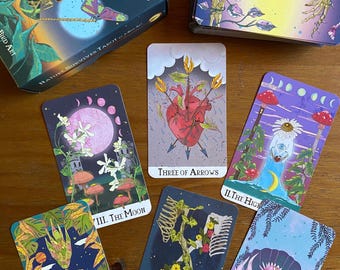 Puede incluir: Una baraja de tarot con un tema de naturaleza. Las cartas presentan ilustraciones de esqueletos, corazones, lunas y otras imágenes inspiradas en la naturaleza. La caja de la baraja es negra con letras doradas y dice "Nature Survives Tarot".