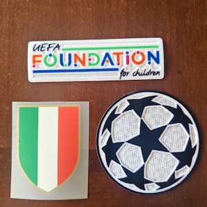 May include: Three patches on a brown surface. The top patch is a white rectangle with the text "UEFA FOUNDATION for children" in colourful letters. Below is a shield with the Italian flag. The last patch is a football.
