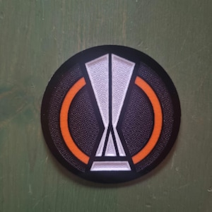 May include: A black and orange circular patch with a white, stylized symbol in the center. The symbol resembles a stylized letter V with a circle around it.