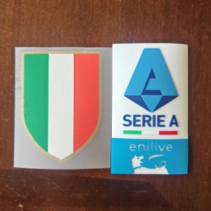 May include: Two stickers, one with the Italian flag in a shield shape and the other with a blue geometric logo and the text "SERIE A enilive".