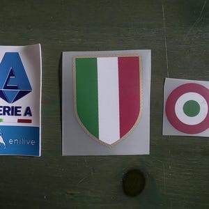 May include: Three rectangular and shield-shaped stickers. The left sticker features a blue Serie A logo with the text "SERIE A" and "enilive" with a dog icon. The center sticker is a shield with the Italian flag colors. The right sticker is a circular badge.