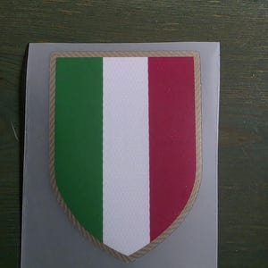 May include: A shield-shaped patch featuring the vertical tricolor flag of Italy with green, white, and red stripes. The design is bordered by a thin, textured tan-colored edge and is set against a plain, light gray rectangular backing sheet.