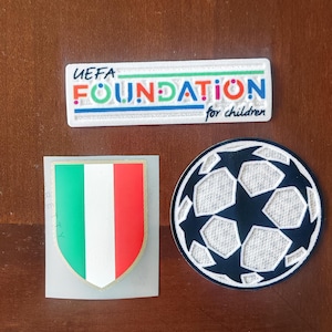 Könnte beinhalten: Drei gestickte Aufnäher: ein weißer Aufnäher mit dem Text "UEFA FOUNDATION for children", ein italienischer Fähnchen-Aufnäher in Grün, Weiß und Rot und ein schwarz-weißer Fußballaufnäher.