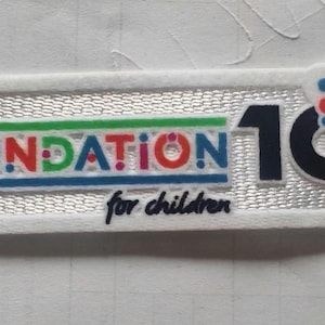 Puede incluir: Un parche rectangular blanco con el texto "UEFA FOUNDATION for children" en varios colores. El número "10" es en negro, con "YEARS" al lado, y un diseño en forma de nube con puntos coloridos.