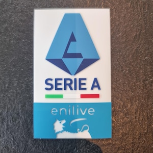 Könnte beinhalten: Ein blauer und weißer Aufkleber mit dem Text "SERIE A" und der italienischen Flagge. Der Aufkleber zeigt auch ein blaues Logo mit einem stilisierten Buchstaben "A" und dem Text "enilive" in Weiß.
