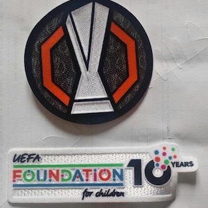 May include: Two patches: a circular one with a black background, orange and white accents, and a trophy design. The second is rectangular, with the text "UEFA FOUNDATION for children" and "10 YEARS" with colourful dots.