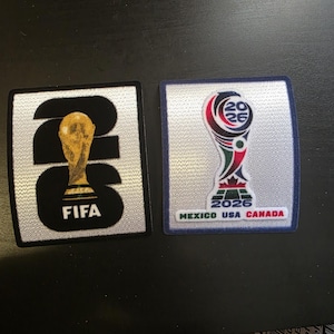 Könnte beinhalten: Zwei rechteckige Aufnäher mit dem Branding der FIFA Fußball-Weltmeisterschaft 2026. Der linke Aufnäher hat einen silbernen Hintergrund mit großen schwarzen Zahlen 26, einer goldenen Trophäengrafik und dem Text FIFA. Rechts das Logo 2026 mit Mexico USA Canada.