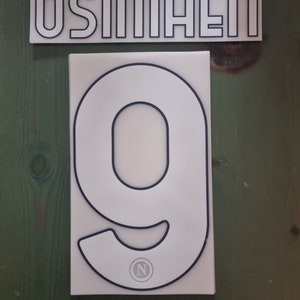Op de afbeelding: Wit en zwart shirtnummer 9 met de naam "OSIMHEN" op een apart wit en zwart naambord. Het nummer heeft een kleine cirkel met de letter "N" erin.