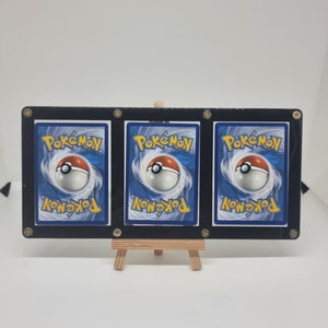 Könnte beinhalten: Drei schwarze Kartenhalter mit transparenten Kunststoffeinsätzen, die jeweils eine Pokemon-Karte mit blau-weißem Hintergrund und einem rot-weißen Pokéball zeigen. Die Halter stehen auf einem kleinen Holzständer.